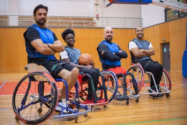 Four athletes in wheelchairs smile confidently, dressed in sports attire, on a basketball court, ready for training or competition.