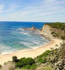 Scenic coastal view featuring a sandy beach, green cliffs, and a rocky outcrop on a clear day with calm blue waters.