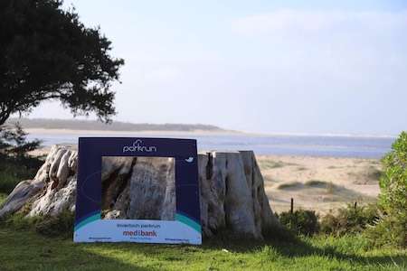 Framed view of a scenic beach with sand dunes and water, featuring a parkrun promotional sign on a tree stump.