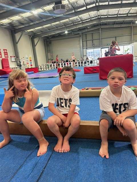 Image displays three young children sitting on a low beam in an indoor gymnastics space. The floor is blue, and two of the children are wearing white with black writing Jets T-shirts.