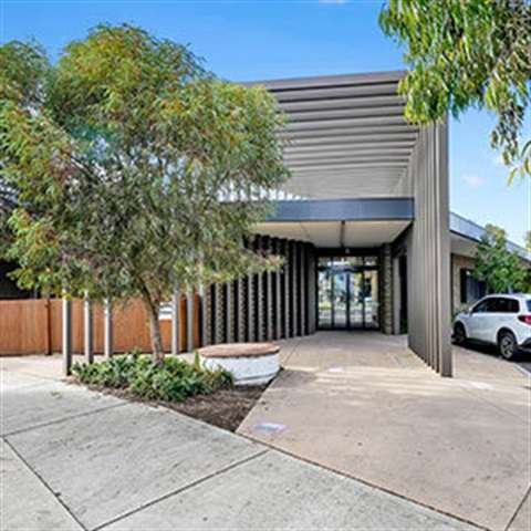 Modern building entrance with a sleek design, featuring glass doors, greenery, and a concrete walkway. Clear blue sky overhead.