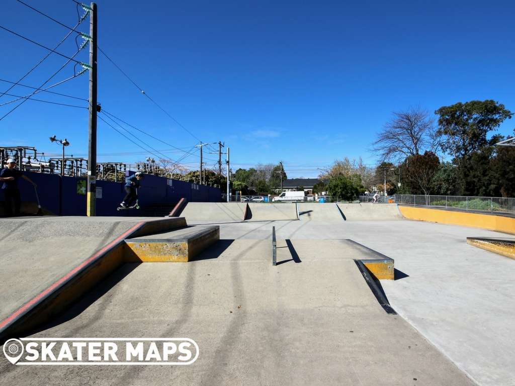 Wide shot of a sunny skatepark featuring ramps and rails, surrounded by trees and a clear blue sky.