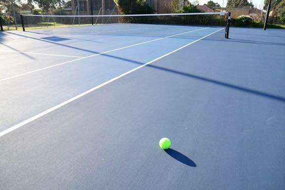 A large tennis court with net with a blue surface and 1 green tennis ball on the ground.