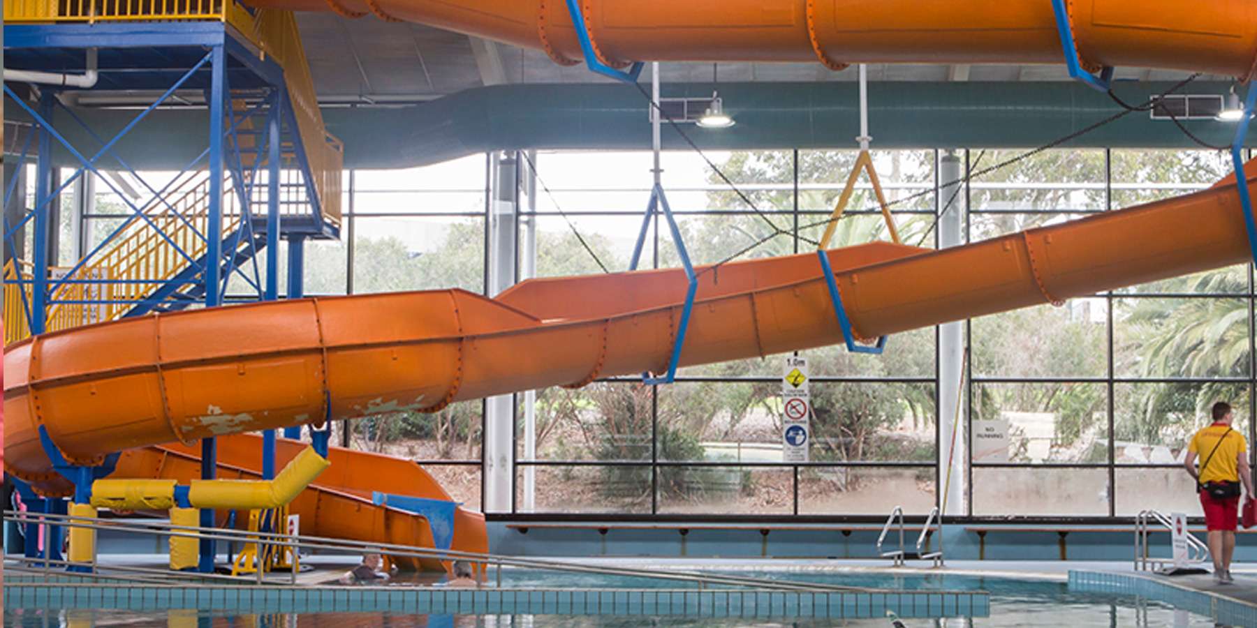 Bright orange water slide curving down into a pool, with blue support structures and a person near the base in a recreational center.