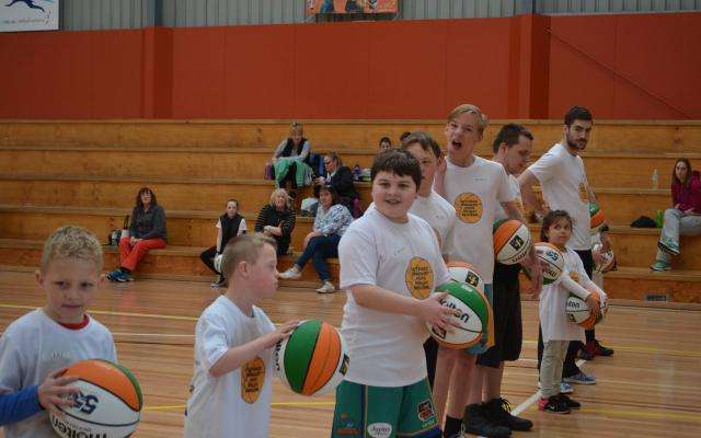 A line of children holding basketballs, smiling, in a gym with spectators in the background, all wearing matching white shirts.
