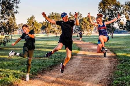 Three individuals joyfully leap on a gravel path surrounded by lush green trees, capturing a moment of fun and fitness outdoors.