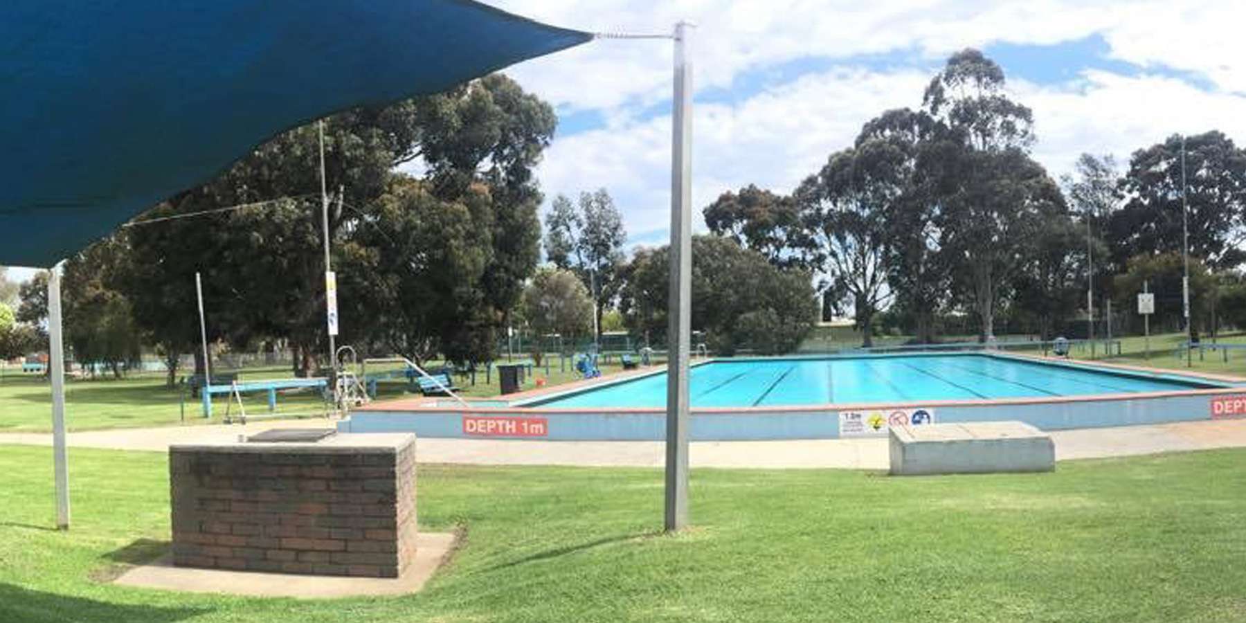 A panoramic view of an outdoor swimming pool surrounded by lush green grass and trees under a partly cloudy sky.