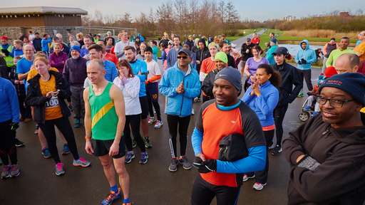 A large group of diverse runners gathers at a park, wearing colorful athletic gear, ready for a race event on a clear day.