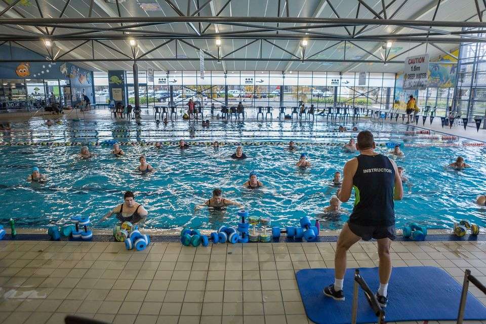 a group of people in a pool doing water aerobics with an instructor standing out side the pool.