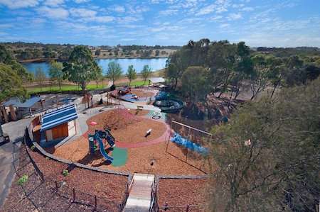 Aerial view of a vibrant playground near a lake, featuring slides, swings, and play structures surrounded by trees and a blue sky.