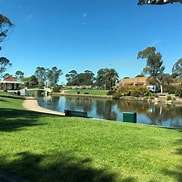 A tranquil park scene featuring a serene lake, lush green grass, trees, and homes under a clear blue sky.