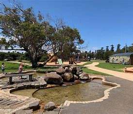 A vibrant park featuring a pond with rocks, a playground, walking paths, and children playing under a clear blue sky.