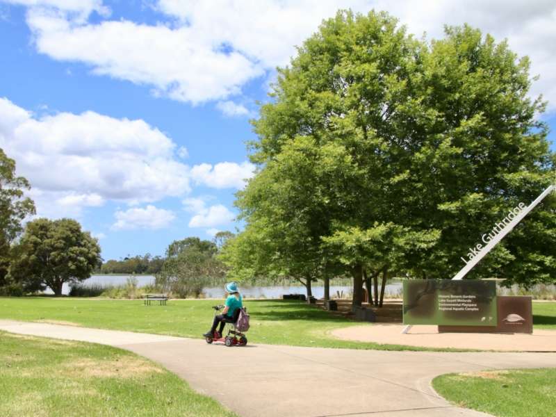 A person on a mobility scooter on a flat path with green grass on each side and a lake in the distance