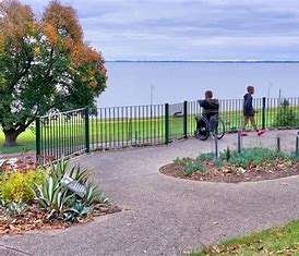 Two people, one in a wheelchair, enjoy a serene lakeside view from a beautifully landscaped path with greenery.