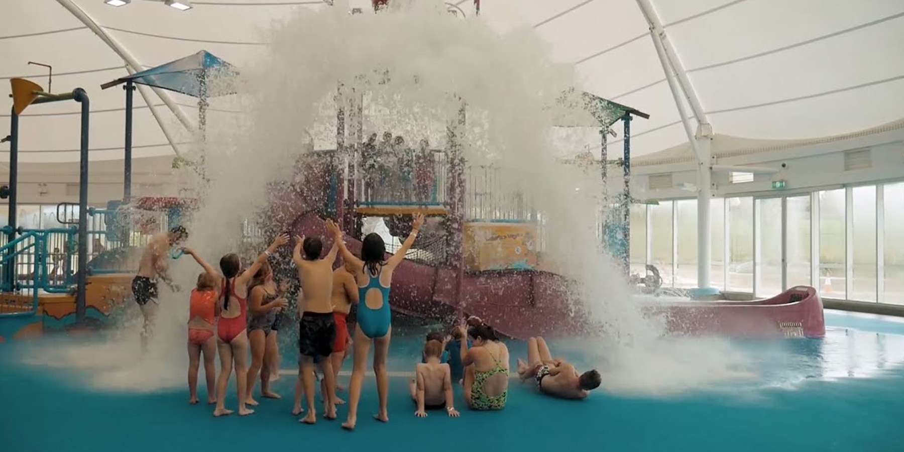 Children in swimwear joyfully gather under a giant water feature, as a torrent of water splashes down in an indoor water park.