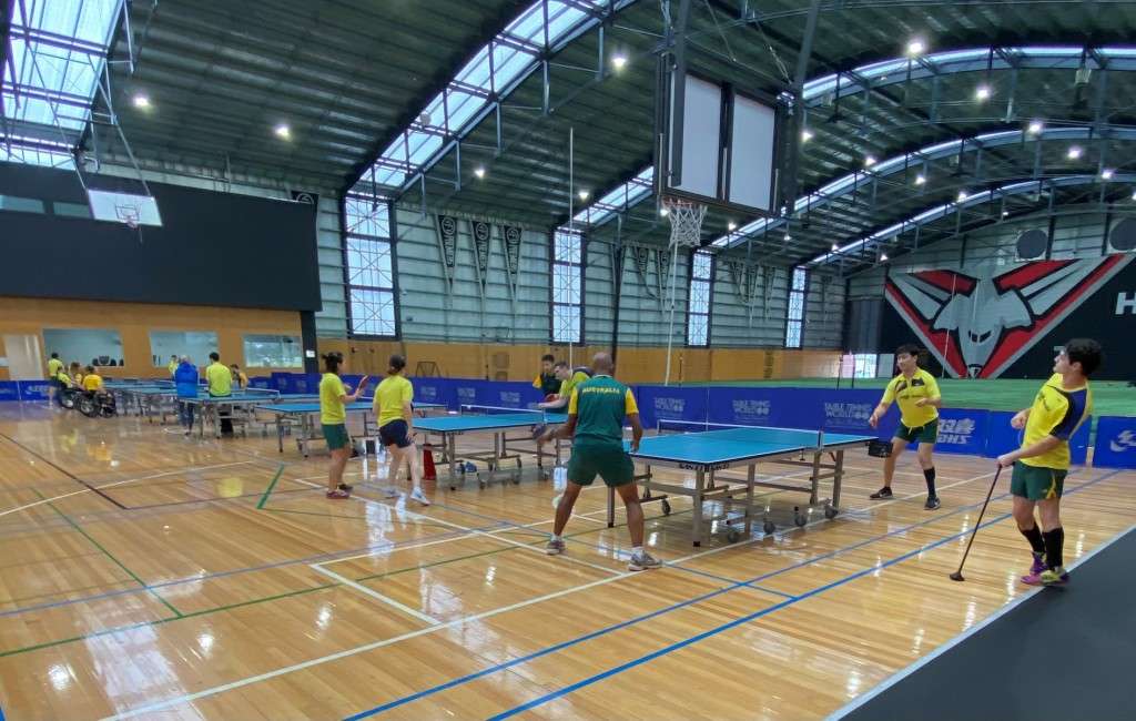 A table tennis training session in a spacious gymnasium with players in yellow and green uniforms practicing at multiple tables.