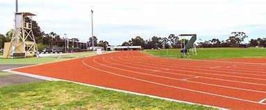 Image displays an athletic track and green reserve grass area, there are green trees in the distant background.