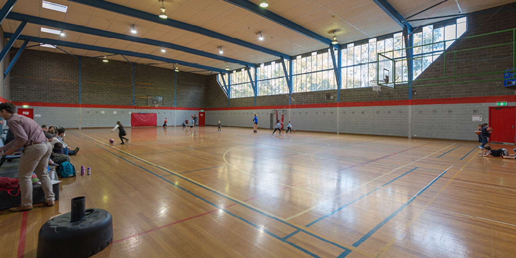 A spacious indoor sports hall with a polished wooden floor, natural light, and players engaged in an activity. Spectators are seated nearby.