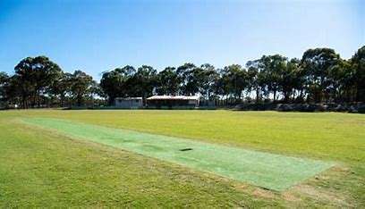 Image displays a green recreation reserve with a cricket pitch, used for sporting and recreation activities. The sky is blue and there are green trees in the background.