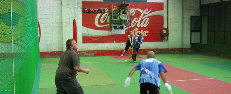 Two players engage in a soccer practice session indoors, with a Coca-Cola sign visible in the background. One player is in goal.