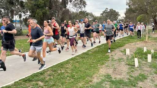 A diverse group of runners jogs along a paved path in a park, surrounded by trees and greenery, under an overcast sky.