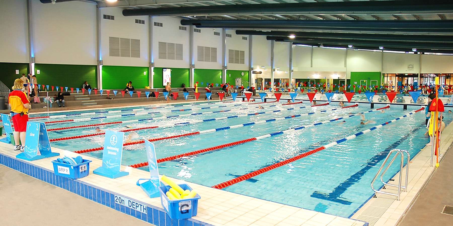 Indoor swimming pool with multiple lanes, lifeguards on duty, colorful walls, and spectators in the background.