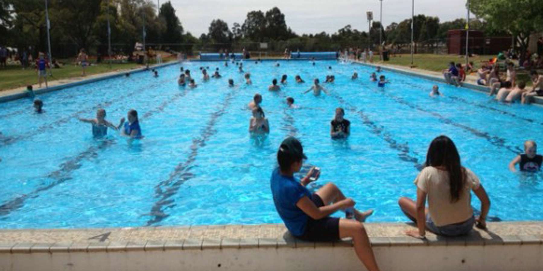 A busy outdoor swimming pool with people swimming and relaxing along the edge on a sunny day, surrounded by greenery.