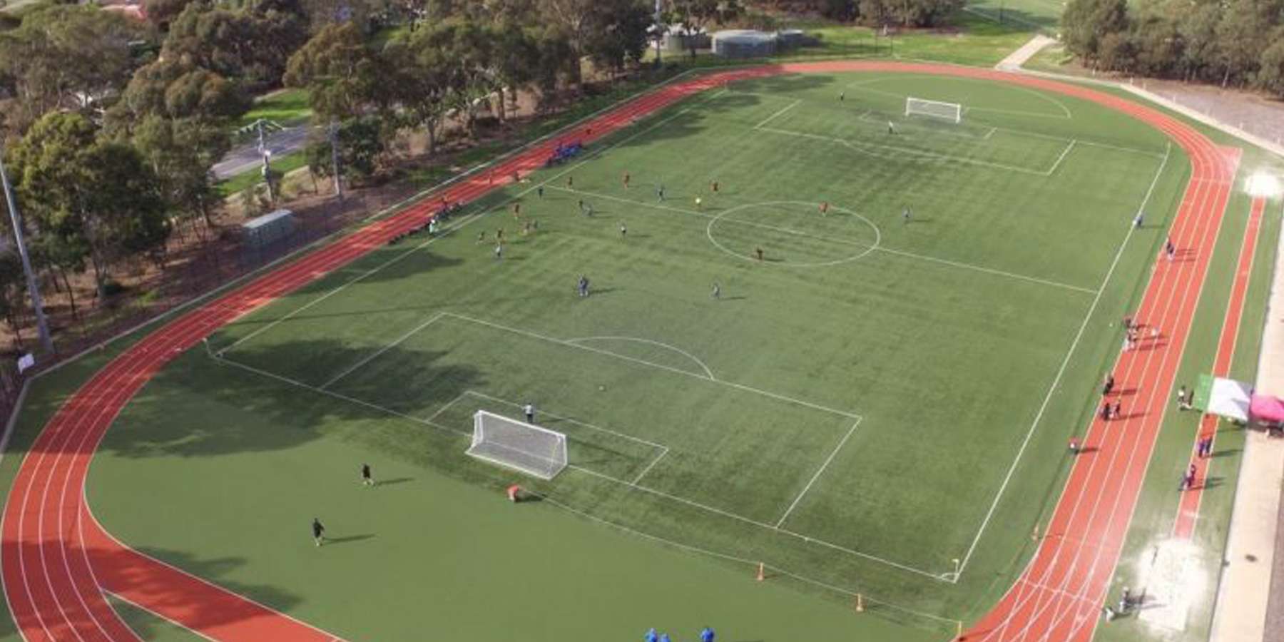 Aerial view of a green sports field with a red track, featuring goalposts and players in action on a sunny day.