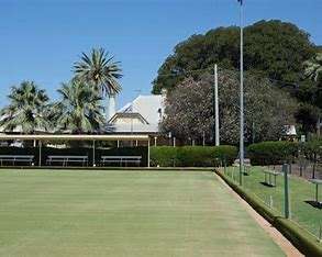 Image displays a flat green croquet ground, with undercover seating in the background. There are palm trees and a blue sky also in the background.