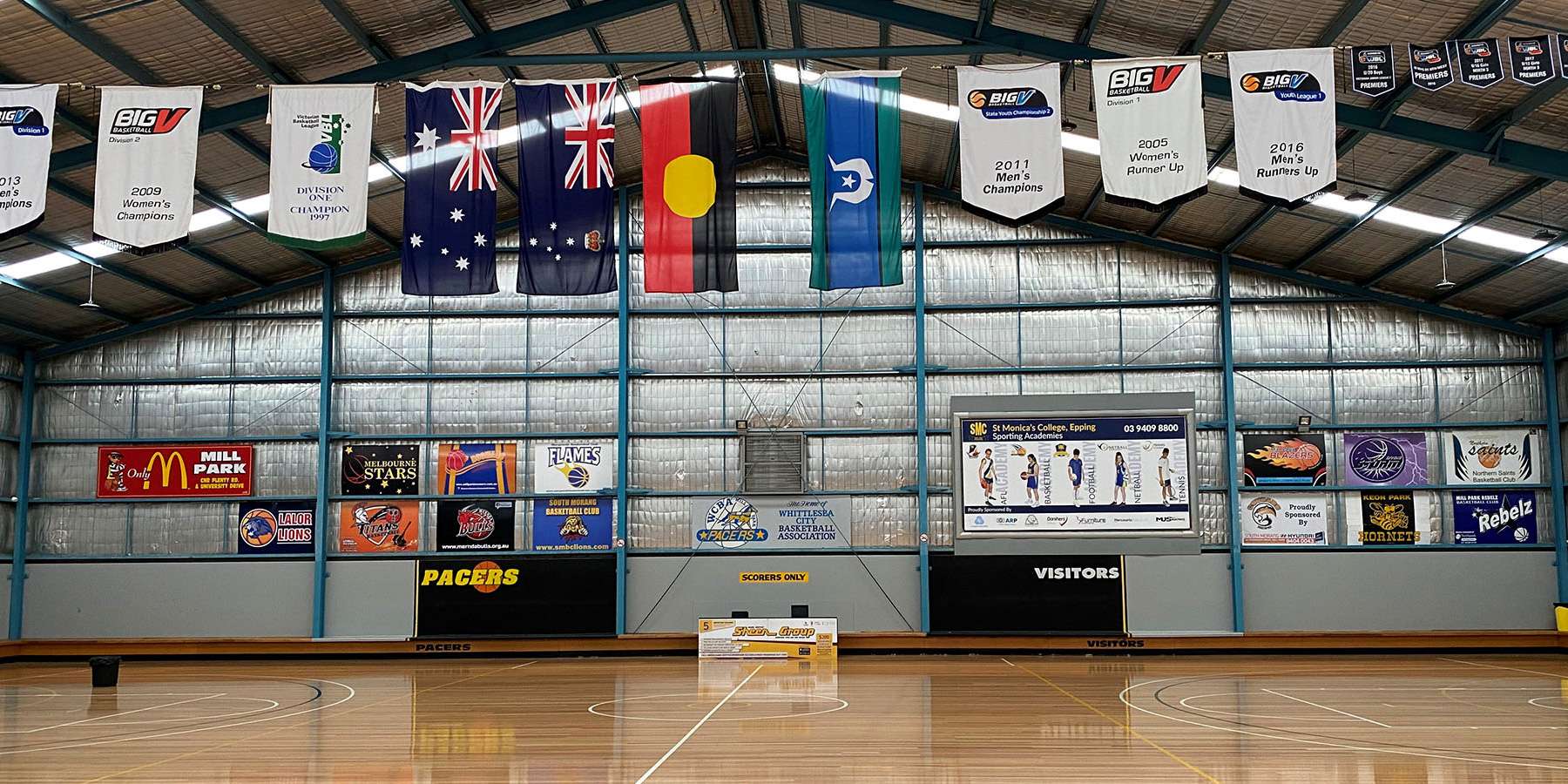 Indoor sports arena with banners hanging from the ceiling, showcasing flags and logos, and a wooden court in front.