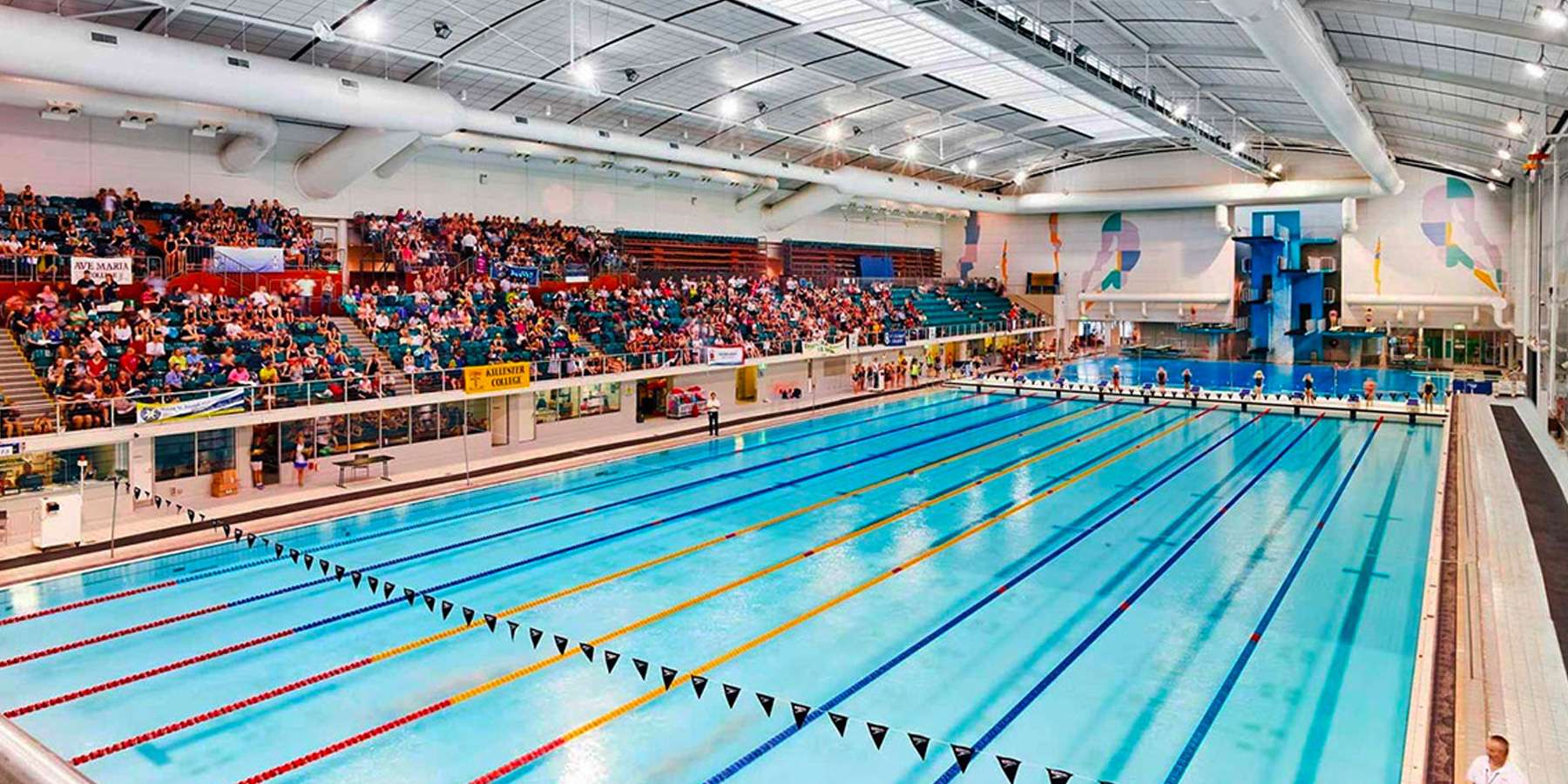 A large indoor swimming pool with multiple lanes, surrounded by a crowded spectator area, under bright overhead lights.