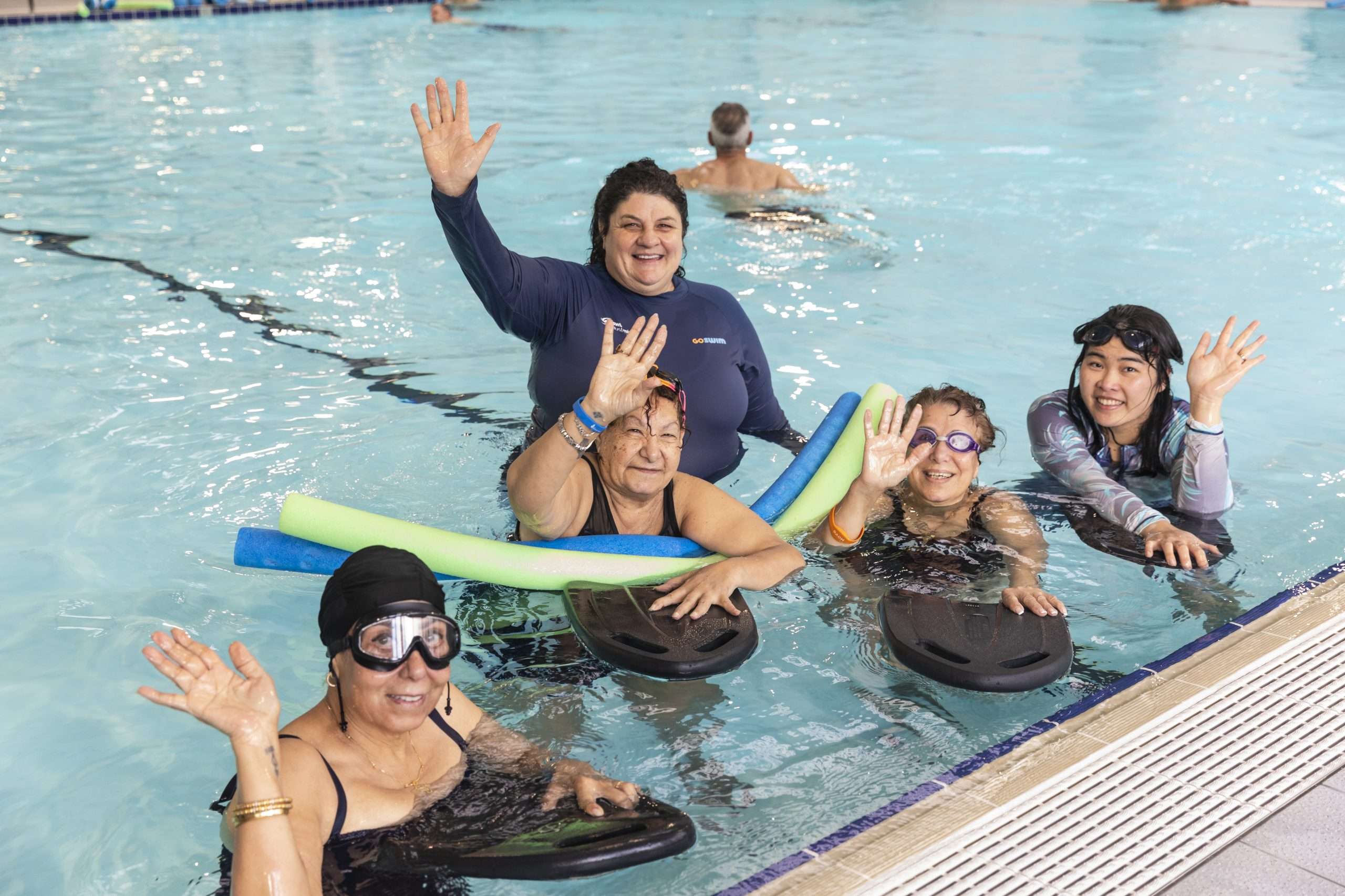 A group of women in swimwear, floating in a pool with pool noodles and float boards, waving and smiling at the camera.