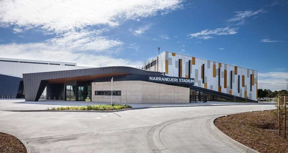 A large white stadium with red abstract roof surrounded by concrete paths and car park.