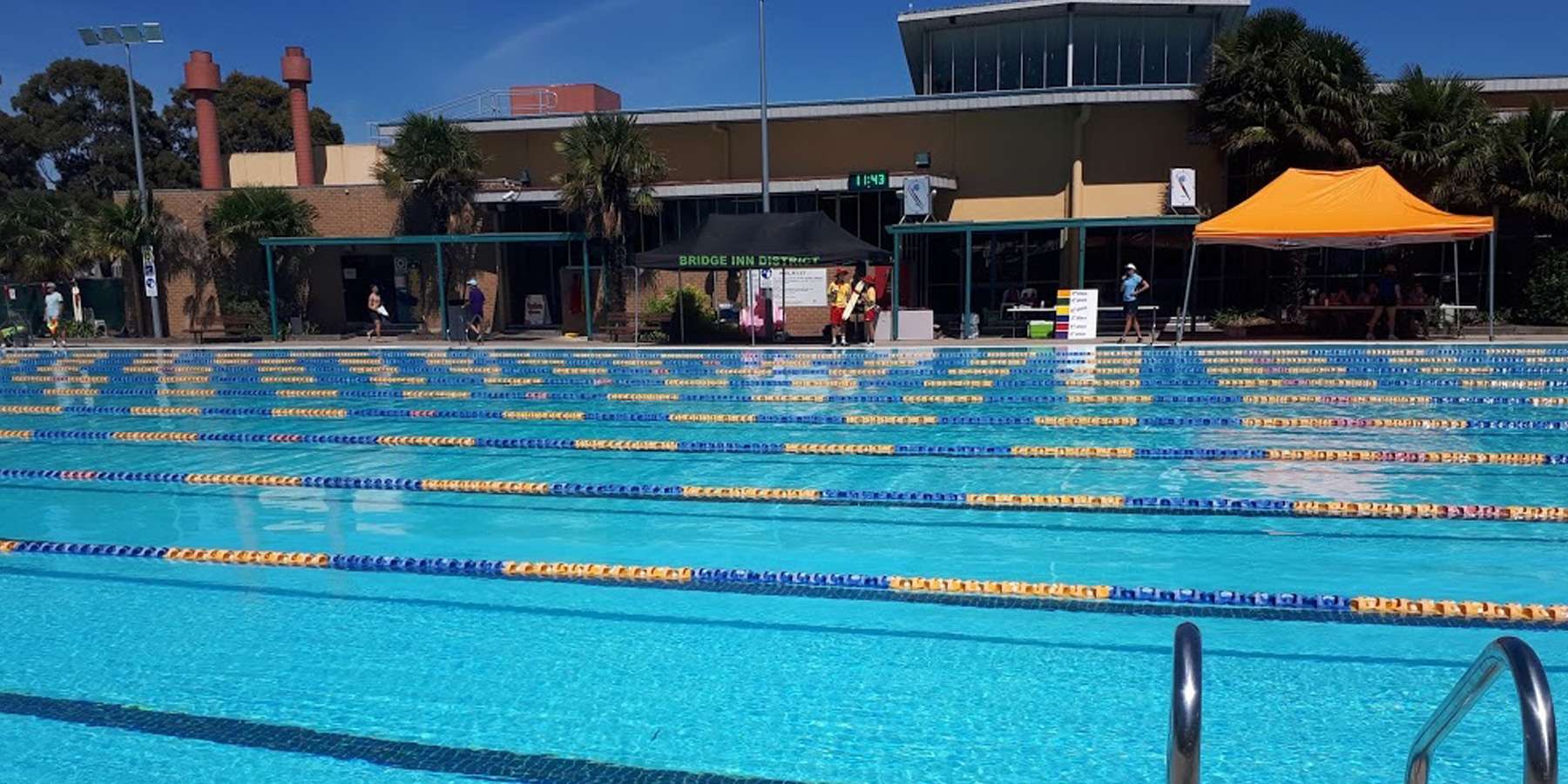 A vibrant outdoor swimming pool with lanes, surrounded by palm trees and a modern building, under a clear blue sky.