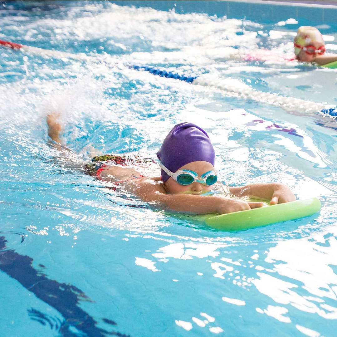 Two children using a green kickboard in the pool, one wearing a purple swim cap.