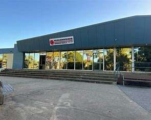 Image displays a concrete path driveway leading to a basketball stadium that has stairs leading up to big glass doors and windows.