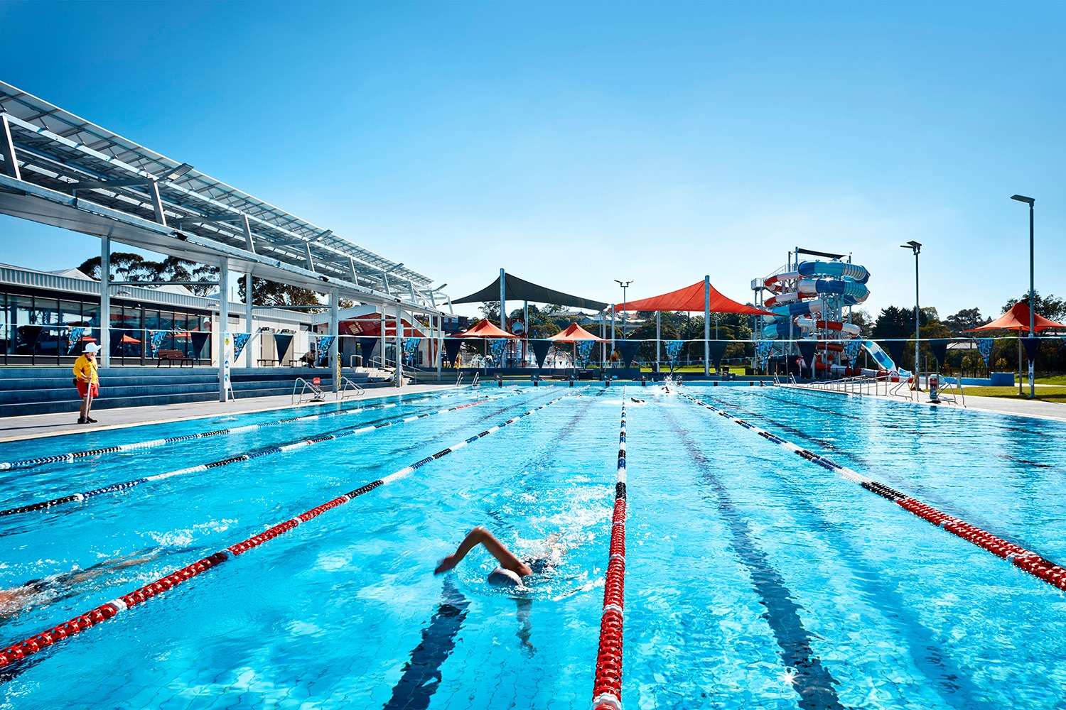 An indoor inground level swimming pool with multiple lanes divided by ropes with red and blue shade sails and red and blue striped water slides.