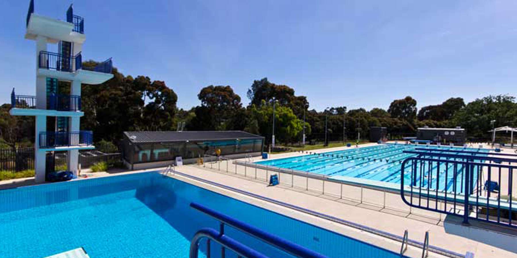 An outdoor swimming pool complex featuring a diving platform, multiple lanes, and surrounded by greenery under a clear blue sky.