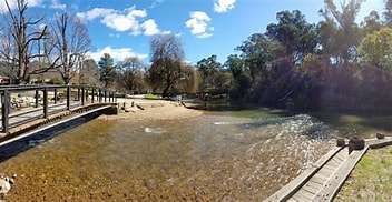 Scenic riverside view with a wooden walkway, clear water, sandy shore, and trees under a sunny blue sky with scattered clouds.