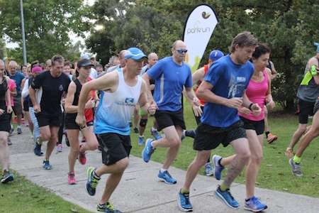 A group of runners in athletic gear participate in a park run, with trees and a white banner in the background.
