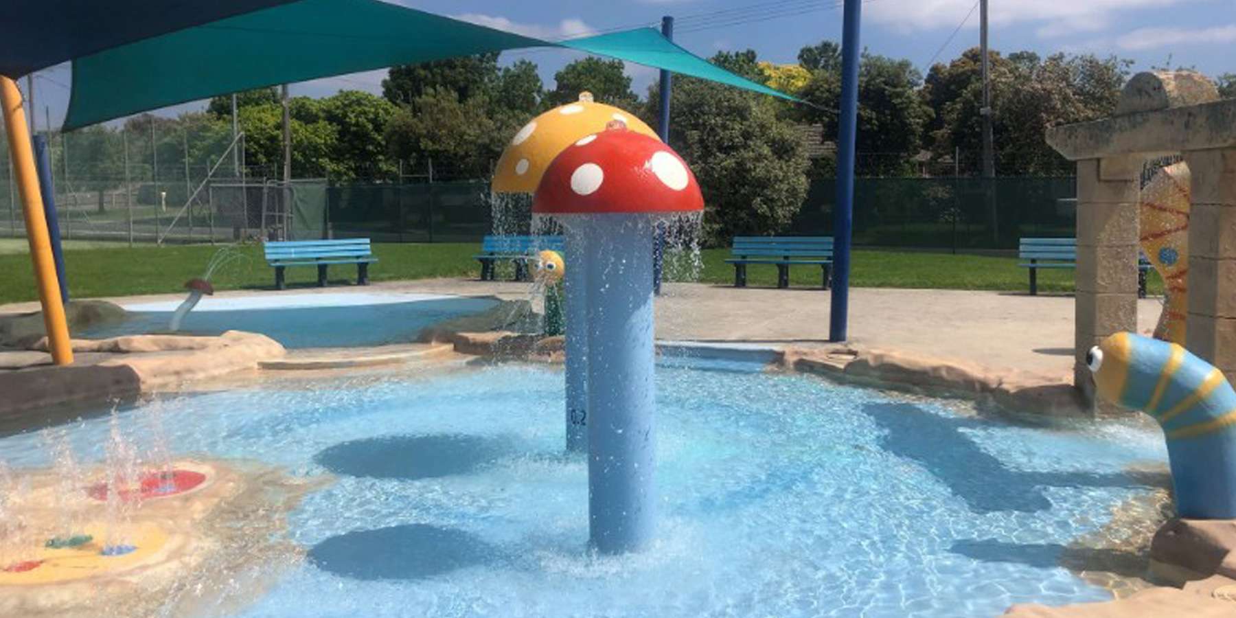 Colorful water play structure with a mushroom cap, spraying water in a sunny park with green benches and shaded areas.