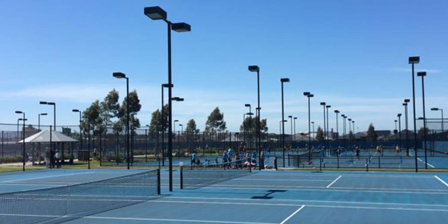A wide view of a bright blue tennis court complex with multiple courts and tall lights under a clear blue sky.