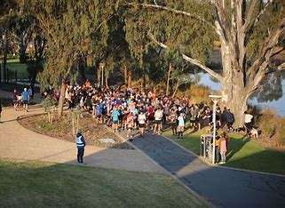 A large group of runners gathers for an outdoor event near a lake. The scene shows participants in various athletic outfits, with some standing near a park pathway. Tall trees provide a natural backdrop, and the sun casts long shadows, creating a warm, evening atmosphere.