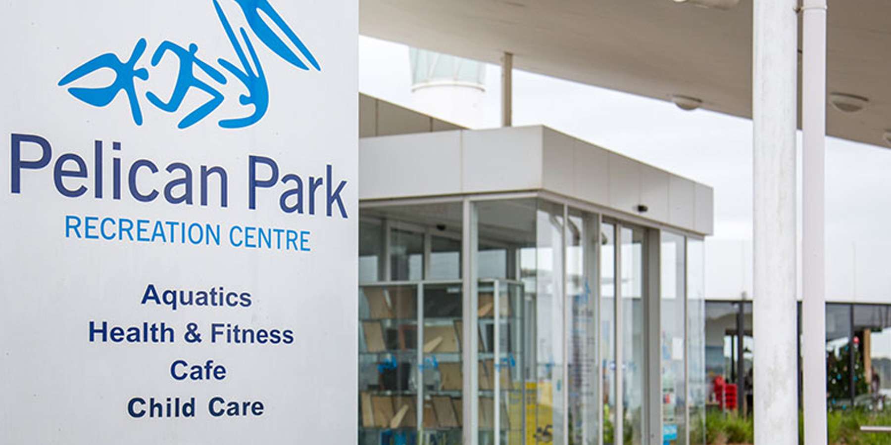 Signage for Pelican Park Recreation Centre, featuring facilities for aquatics, fitness, cafe, and child care. Modern building entrance visible.