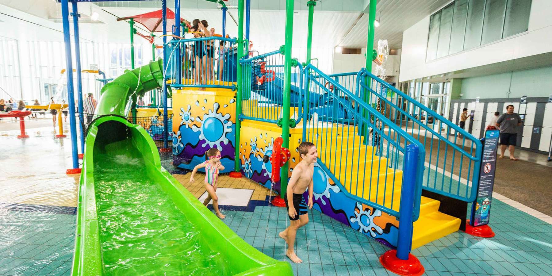 Children playing at a colorful indoor water slide complex with stairs, vibrant artwork, and a green slide.