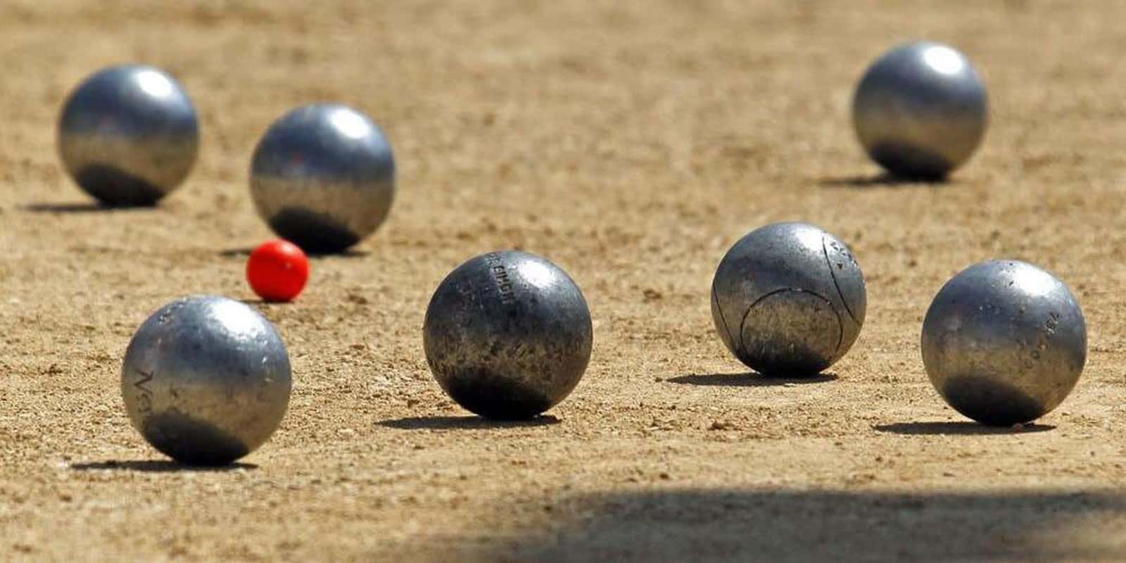 A game of pétanque featuring six metallic balls scattered on a sandy surface, with a small red ball positioned among them.