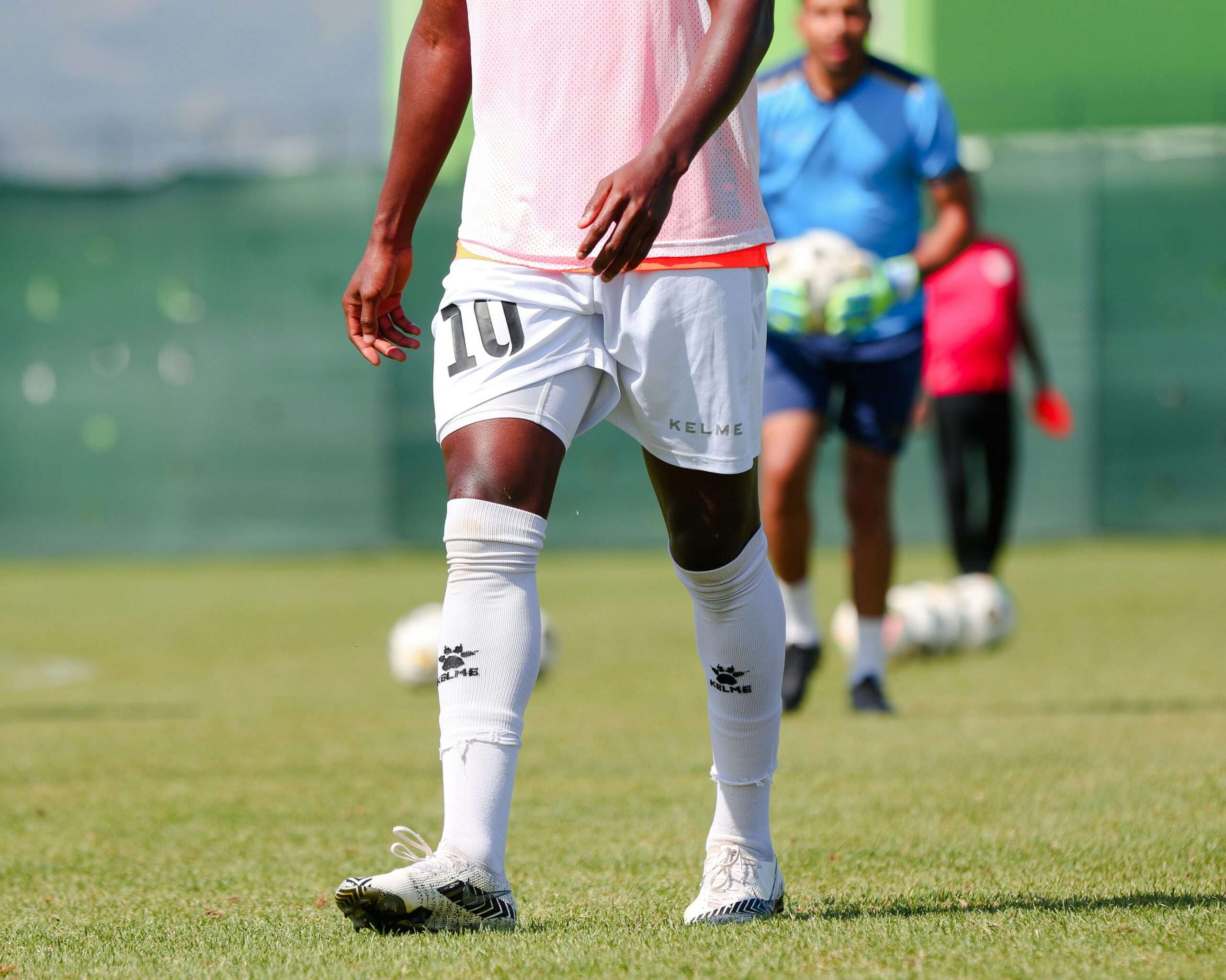 A soccer player in a pink jersey and white shorts walks on the field, with coaches and players practicing in the background.