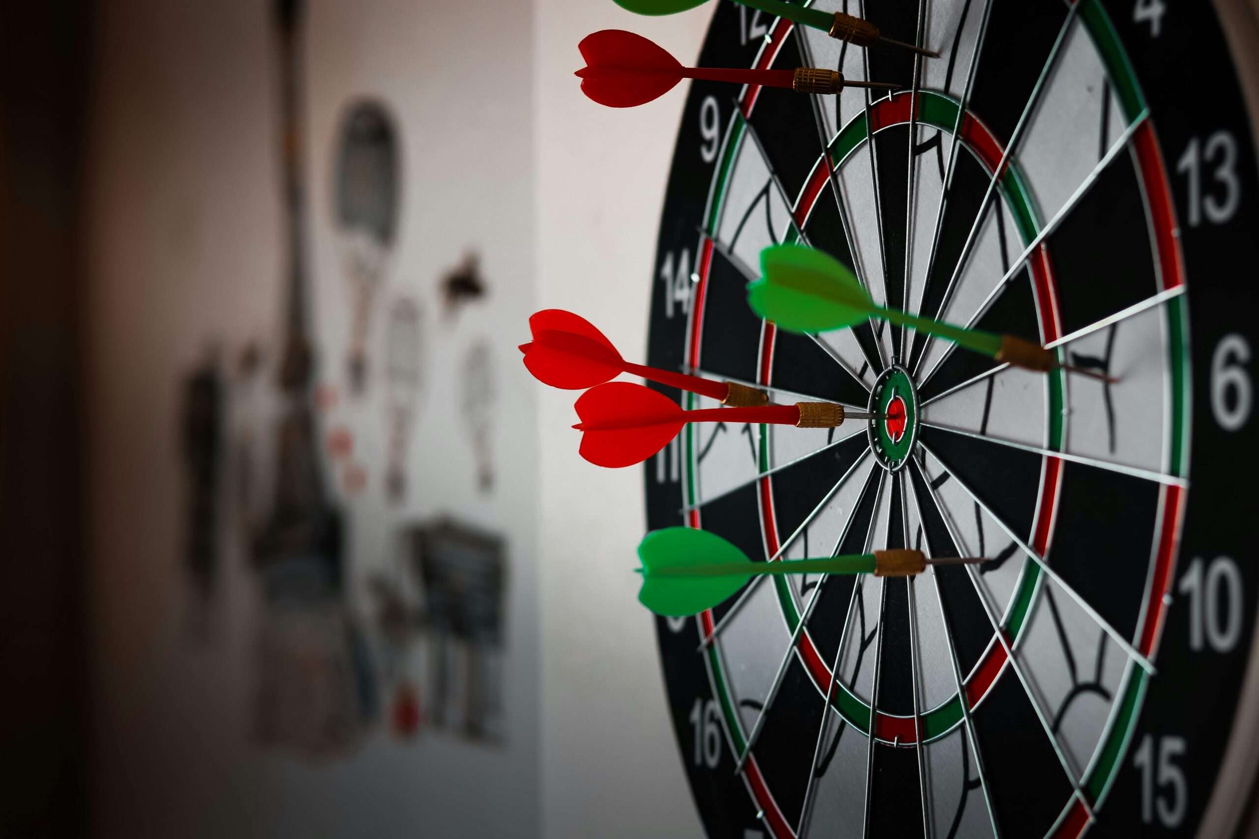 Close-up of a dartboard displaying several red and green darts embedded in its target, with blurred illustrations in the background.