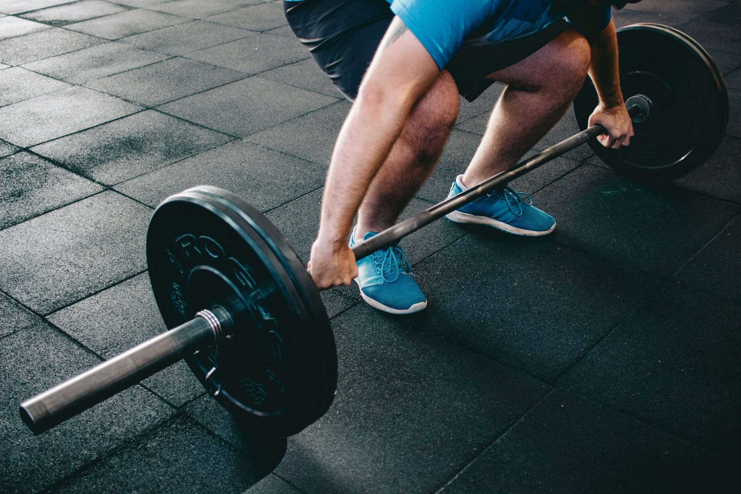 A person in blue shorts and sneakers prepares to lift a barbell on a gym floor, emphasizing strength training.