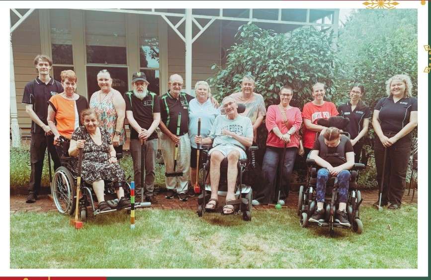 A group photo of individuals, some in wheelchairs, posing outdoors in front of a house and greenery. Smiles and diversity are evident.
